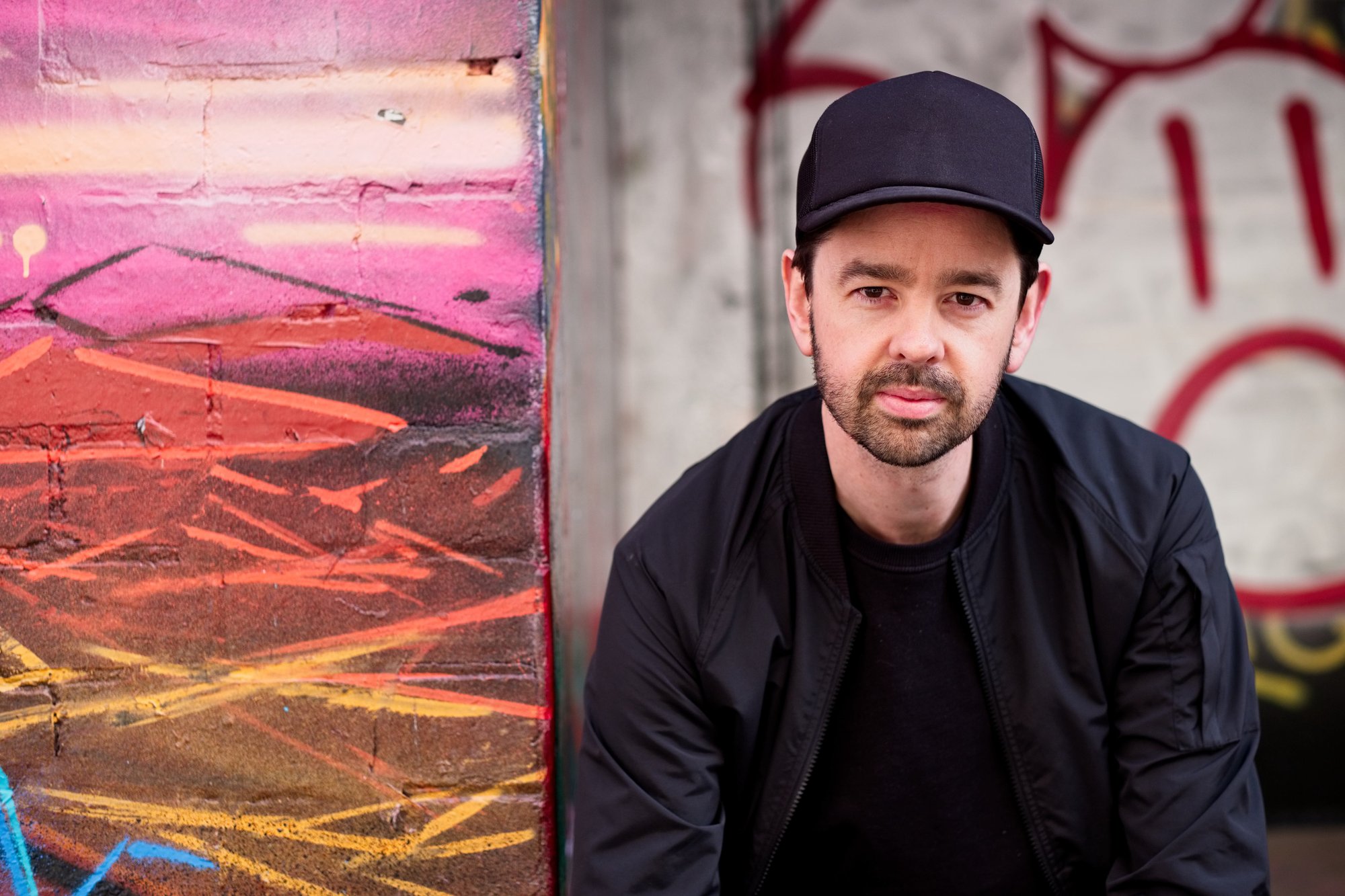 Rob Ward in a black cap and jacket beside a painted graffiti wall